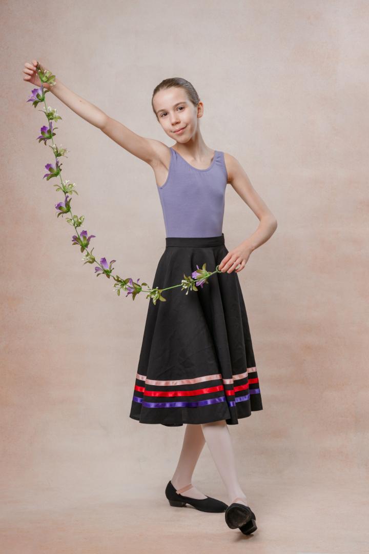 A young ballet student holding a flower garland while dressed in a lavender top and black skirt.